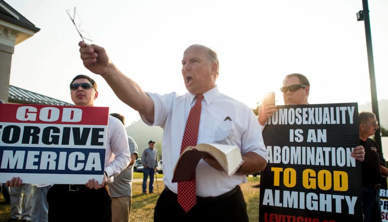 person in tie shouts holding book; people on either side hold signs that say "God Forgive America" and "Homosexuality is an abomination to God almighty" (zero-sum beliefs)