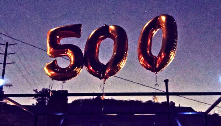 Balloons that read "500" float in the sky while tied to a pole at dusk