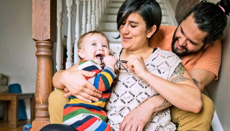 parents and baby sit at foot of stairs, baby tries to eat glasses