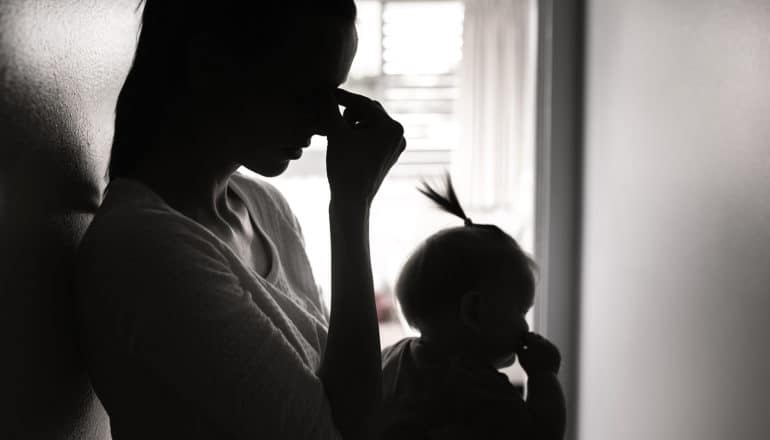 A mother touches her face while holding her baby in an apartment hallway