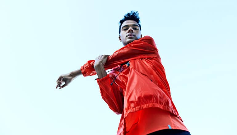 A man in a red windbreaker stretches his arm against a blue sky