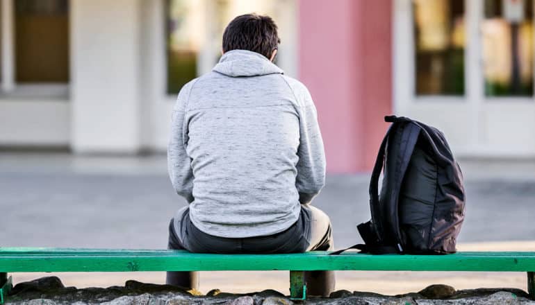 A teen boy sits alone on a green bench next to a black backpack