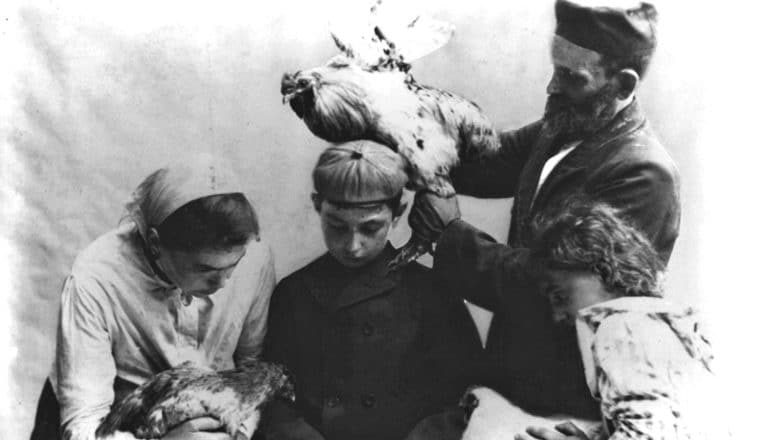 A family sits at a table while the father holds a chicken over their head