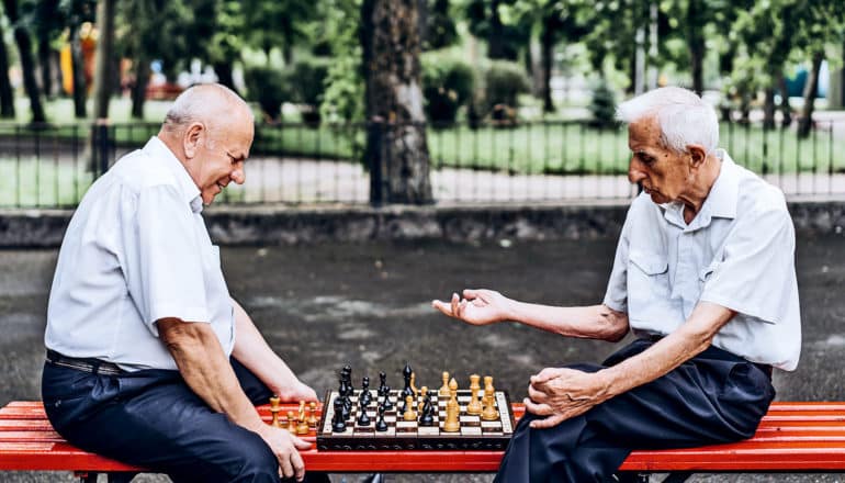 Two older men sit on a red bench in a city park playing chess