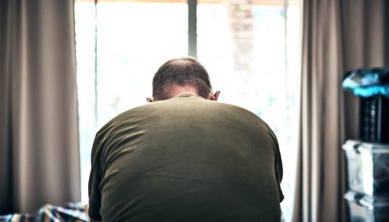 A man sits on his bed looking down facing away from the camera