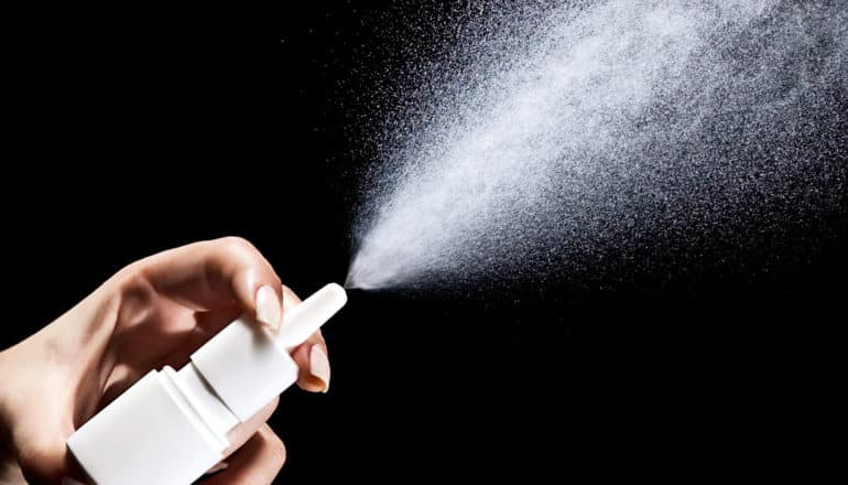 A person sprays a bottle of nasal spray against a black background