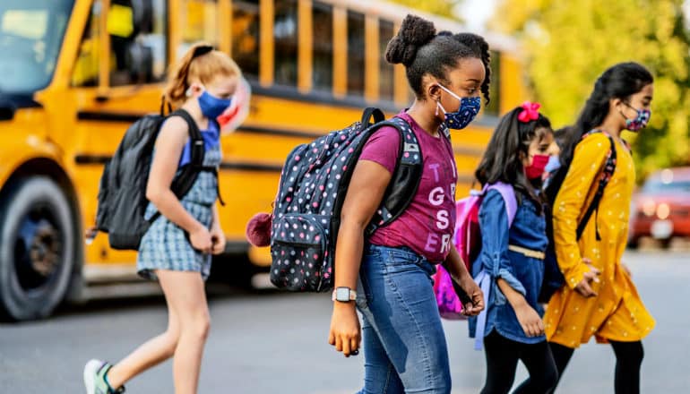 Kids in masks and carrying backpacks walk from a school bus