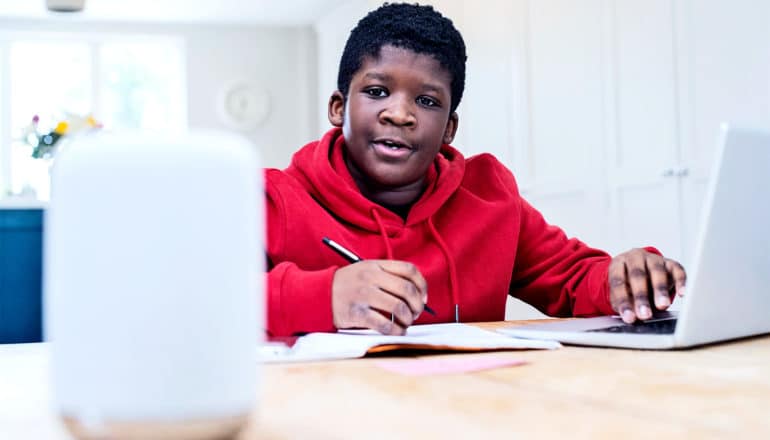 A young boy in a red hoodie does homework at a laptop while talking to a voice assistant device