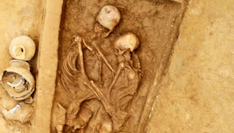 Two skeletons buried together embrace in a coffin buried in yellow dirt