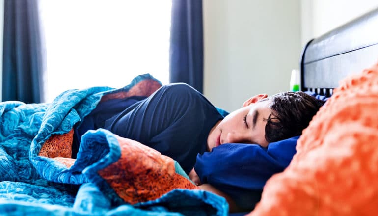 A teen boy sleeps in a bed with blue and orange blankets while light comes in through the window