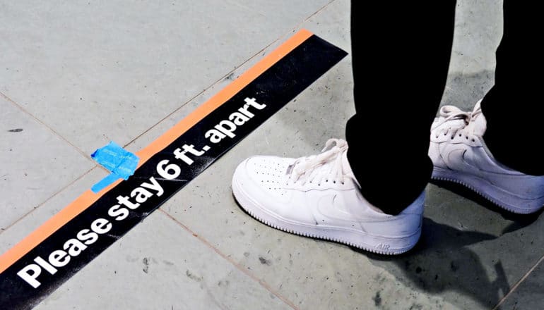 A person in black pants wearing white shoes stands at a sign on the floor that reads "Please stay 6 ft. apart"