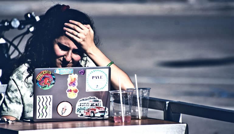 A young woman works on a laptop outside at a cafe while resting her head on her hand