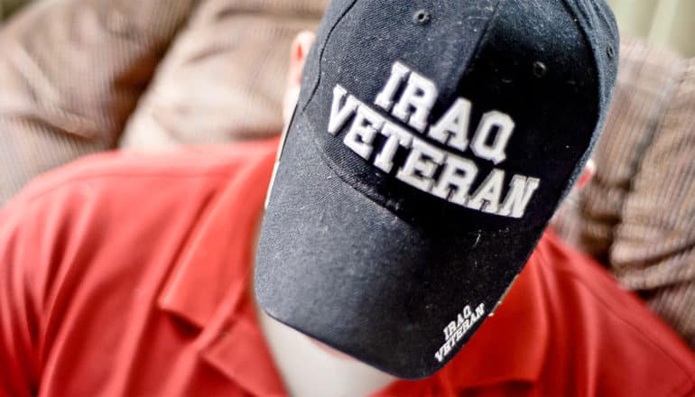 A man looks down while wearing a baseball cap that reads "Iraq Veteran" and sitting on a couch