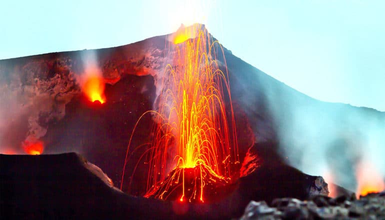 A volcano erupting shoots arcs of lava and sparks while smoke rises into the blue sky