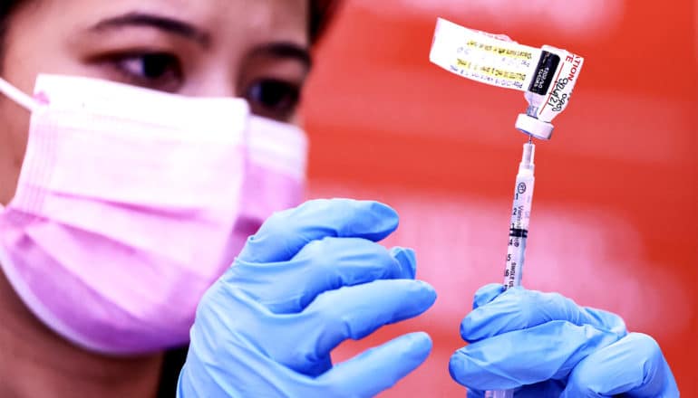 A nurse fills a syringe with the covid-19 vaccine while wearing a pink face mask and blue gloves