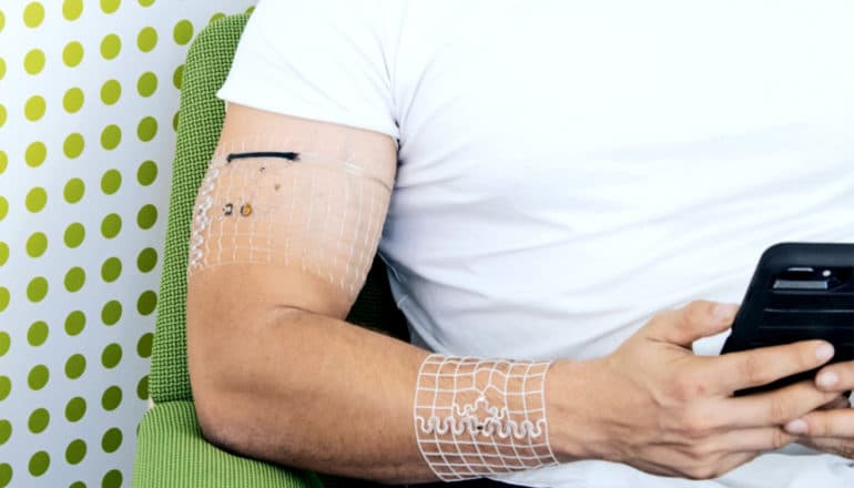 A man sits in a chair looking at his phone with the clear plastic wearable devices around his wrist and upper arm