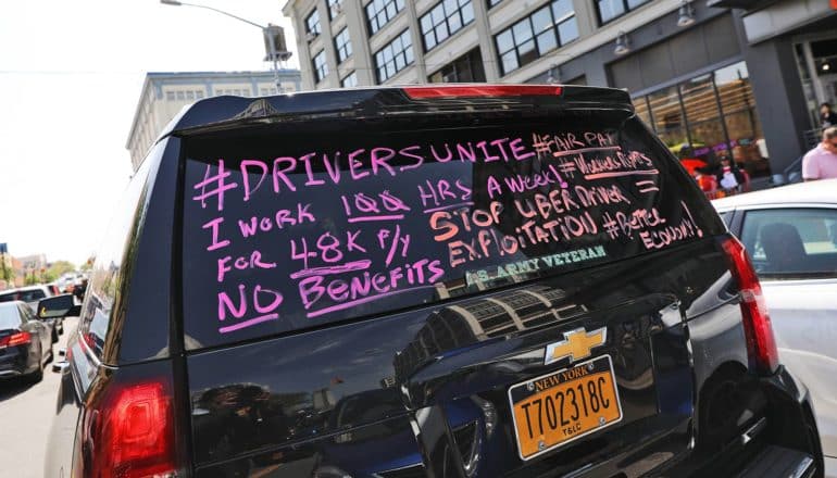 writing on vehicle back window: "#DriversUnite #FairPay I work 100 hrs a week for 48K/yr Stop Uber driver exploitation No benefits #workersrights #bettereconomy"