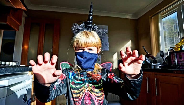 A young girl has a black skeleton shirt, fairy wings painted black, a black unicorn horn, and a COVID face mask on