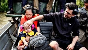 A mother holds a baby as the baby's father touches his forehead. The couple and baby are seated in a rescue boat