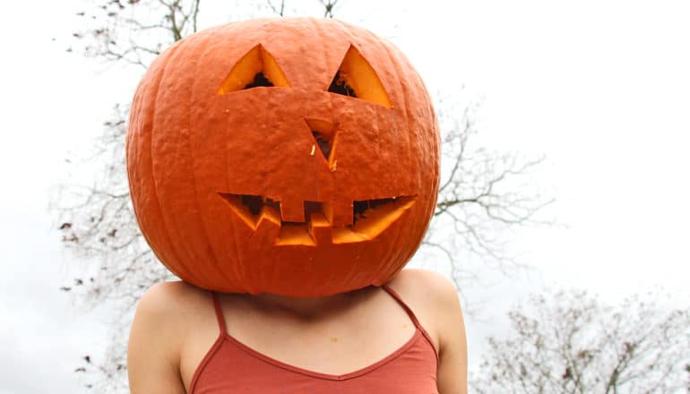 A woman wears a pumpkin on her head with a smiling face carved into it