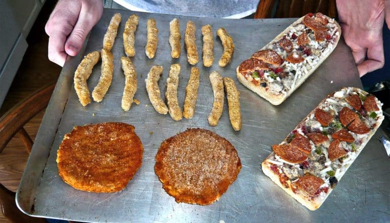 A person holds a baking tray with frozen foods, including two pizza slices, two patties of meet, and mozzarella sticks