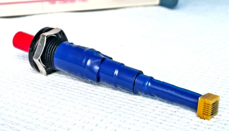 The device is a long blue tube with an orange section with microneedles on one end and a black and red top on the other