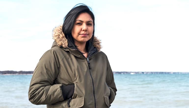 An indigenous woman walks along a beach in a winter coat