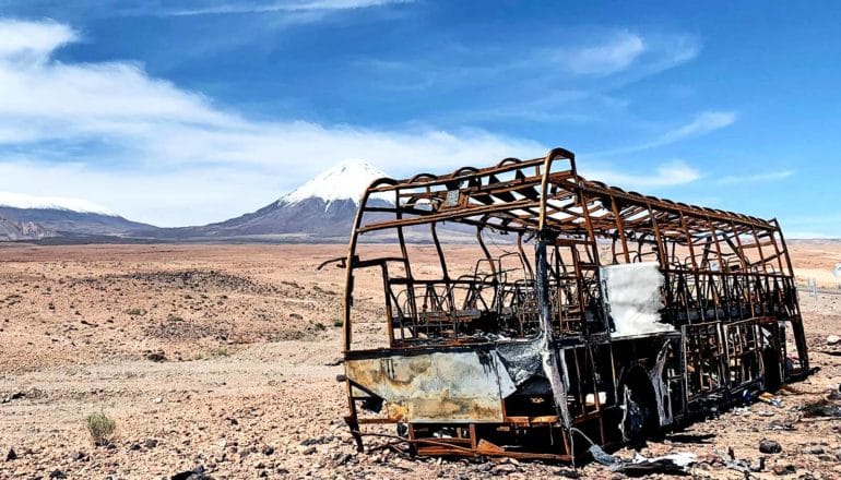 A rusted bus frame sits in the Atacama desert with a huge white-capped mountain in the distance