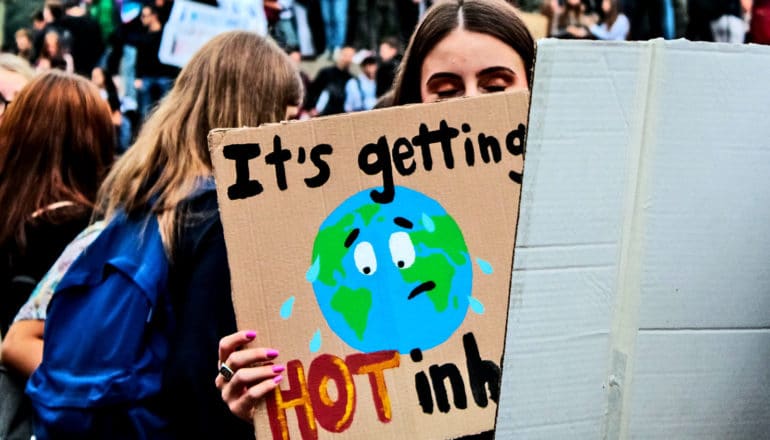 A young woman at a protest for action on climate change holds a sign that reads "It's getting hot in here" and shows a picture of a sweating Earth