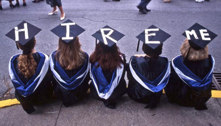 Five college graduates sit on a curb in their caps and gowns. Their caps have the words "Hire me" spelled out in white tape