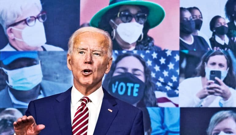 Joe Biden speaks in front of a screen showing people in COVID-19 face masks