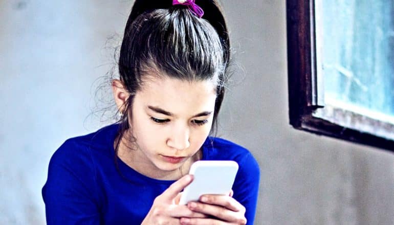 A young teen girl holds a smartphone and looks at the screen while sitting in a stairwell