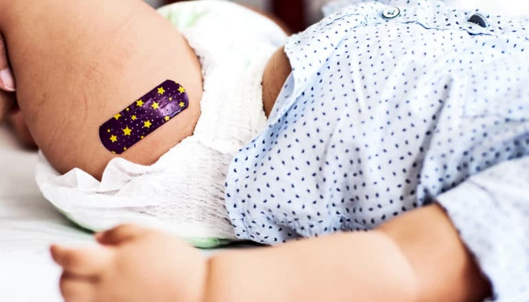 A baby in a diaper and blue shirt has a bandage over a vaccination wound on their thigh