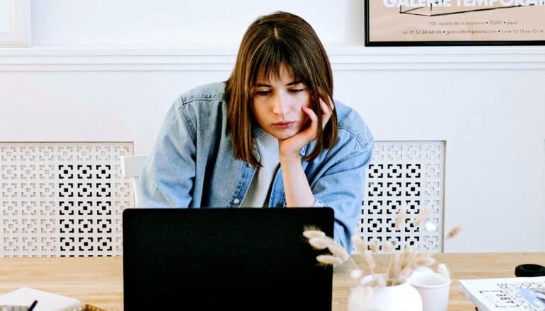 A young woman sits resting her chin on her hand while sitting at her desk looking at her laptop