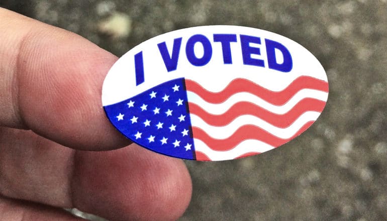 A person holds up an "I Voted" sticker with the American flag on it