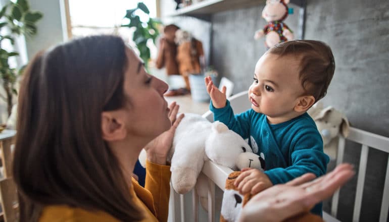 A baby standing in a crib looks like he's speaking to his mother, who is speaking back and gesturing with her hands