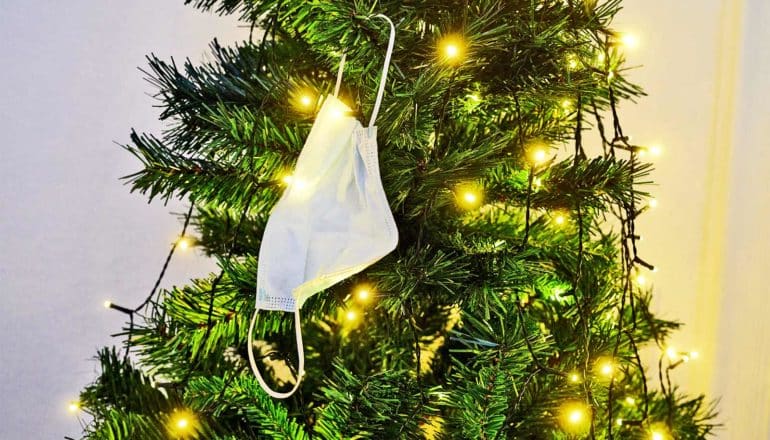 A medical face mask hangs from a green Christmas tree covered in yellow lights