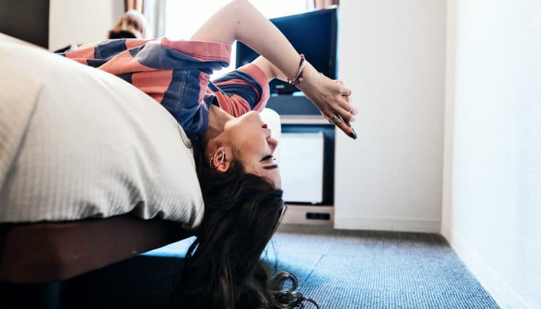 A young woman is laying on her back on the edge of her bed while using her phone upside down