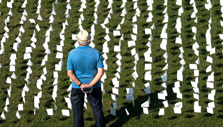 Hundreds of white flags on grass in front of a man in a blue shirt standing with his hands clasped behind him