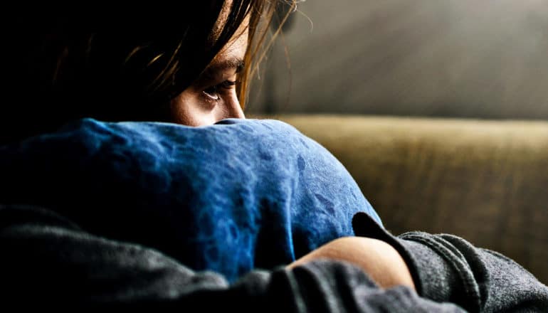 A teen hugs a blue pillow to her face while sitting on a couch