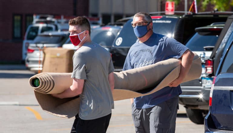 A father and son carry a rolled up rug in a college dorm parking lot