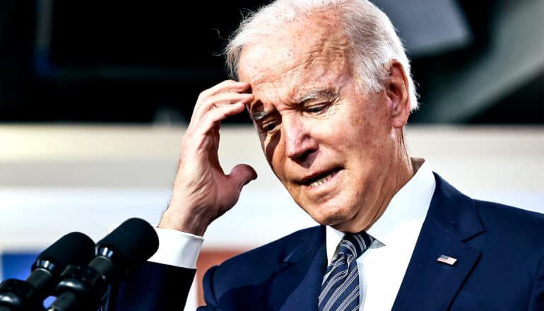 President Joe Biden touches his forehead while standing at a podium speaking into microphones