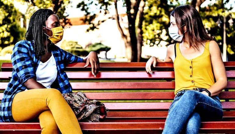 Two women sit in a park talk with masks on