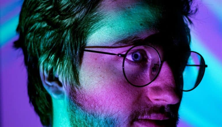 A young man wearing glasses has blue and pink light projected onto his face