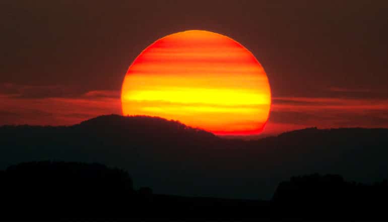 The sun appears bright red and orange as it sets behind dark mountains