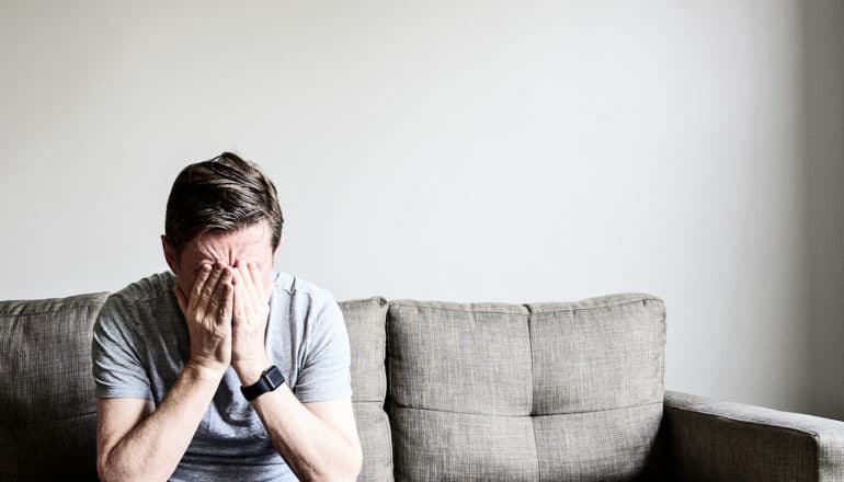 A man sits alone on a couch covering his face with his hands