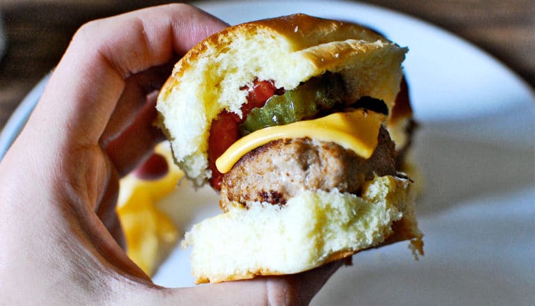 A person holds a small turkey burger with cheese, tomato, and a pickle