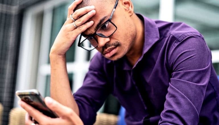 A man looks down at his phone while putting his hand to his forehead