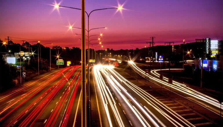 Cars drive on a highway at sunset under a purple sky