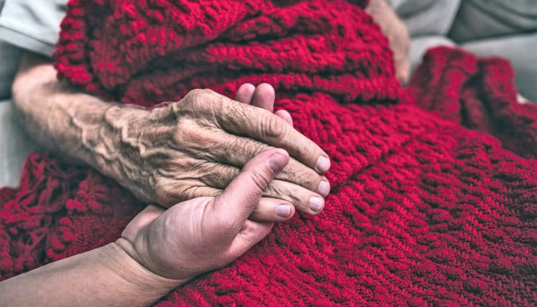 An older patient holds a person's hand while sitting under a red blanket